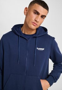 Sweat à zip marine fabriqué en tissu doux, avec un logo sur la poitrine, une capuche à cordons et des poches avant. Texture lisse.