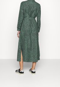 Robe longue à motifs de feuilles vertes avec des manches longues, ceinture à la taille, fente sur le côté, et assortie à des baskets de sport noires et blanches.