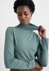 Maglione a maniche lunghe, color teal, con collo a dolcevita in morbido tessuto testurizzato, dal design aderente e collo arrotolato, abbinato a pantaloni oliva a coste.