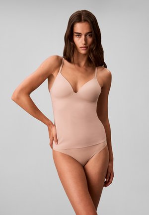 Femme portant un top caraco nude ajusté avec de fines bretelles et une lingerie nude assortie, debout avec une main sur la hanche devant un fond uni.