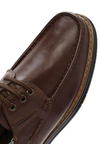 Chaussure en cuir marron avec surface texturée, embout arrondi et couture contrastante. Elle présente des lacets tressés et une semelle en caoutchouc avec un petit talon.