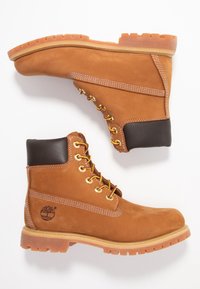 Bottes de travail en cuir marron avec œillets dorés, lacets beige et semelle robuste. Présentent un talon marron foncé contrastant et un logo sur le côté.