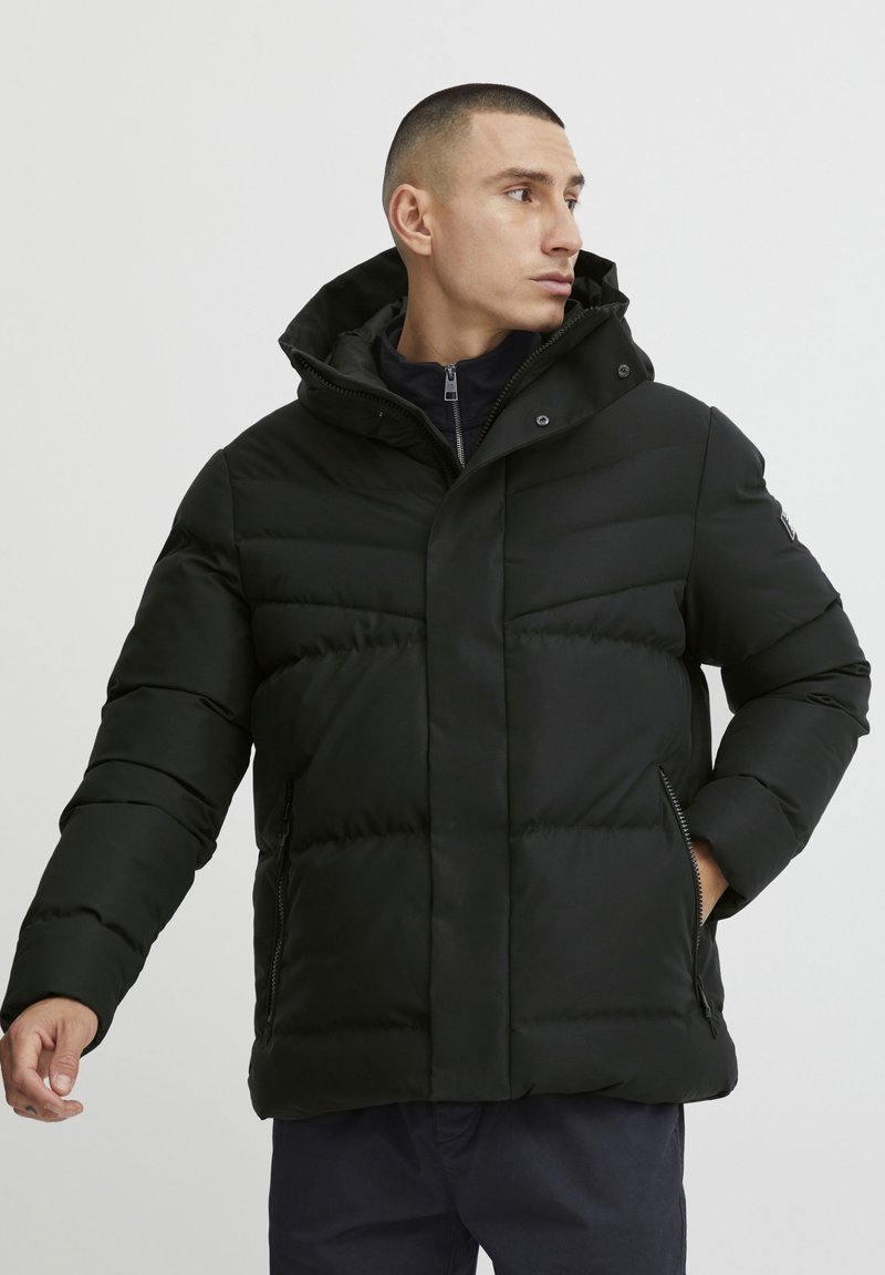 Sort polstret jakke med aftagelig hætte, der har lynlås foran, sidelommer og en quiltet design for varme og isolering.