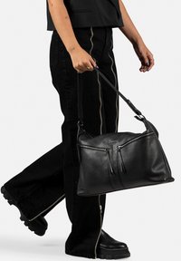 Borsa a spalla in pelle nera con due tasche con zip, finitura opaca, tracolla regolabile e forma rettangolare. Indossata con pantaloni neri e stivali.