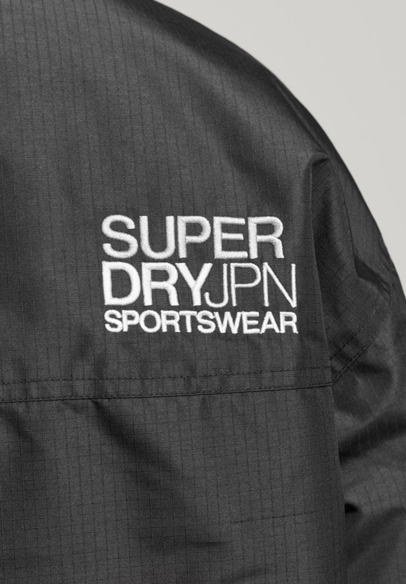 Zwarte sportjack met een gestructureerde stof, voorzien van het geborduurde logo "SUPERDRY JPN SPORTSWEAR" in het wit op de linker schouder.