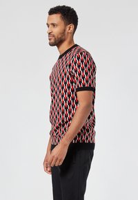 Suéter de punto de manga corta con un patrón geométrico en rojo, blanco y negro, con un cuello y puños de canalé. Diseño estructurado y ajustado.