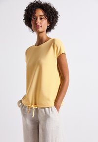 Žlutý top s krátkým rukávem a kulatým výstřihem, texturovaný, s nastavitelnou tkaničkou na spodním okraji, kombinovaný s světlými lněnými kalhotami.