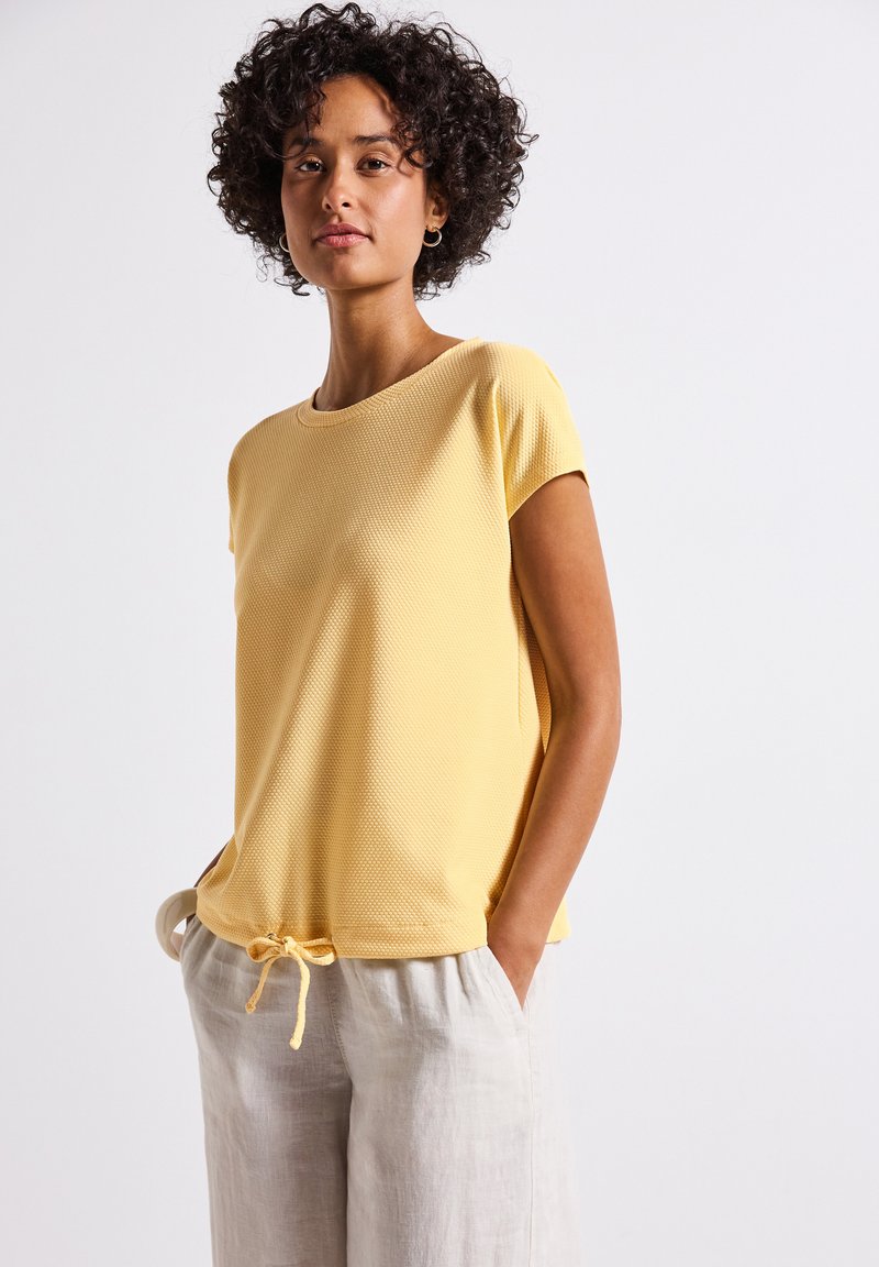 Žlutý top s krátkým rukávem a kulatým výstřihem, texturovaný, s nastavitelnou tkaničkou na spodním okraji, kombinovaný s světlými lněnými kalhotami.