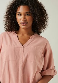 Vaaleanpunainen lyhyttakkinen paita, jossa on V-kaula-aukko, kaksi rintataskua ja käärityt hihat. Valmistettu kevyestä kankaasta, jossa on sileä rakenne.