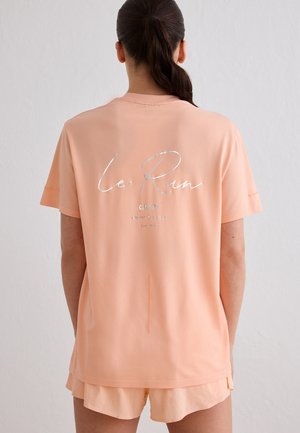 Kvinde med mørk hestehale iført ferskenfarvede shorts og matchende oversized T-shirt med sølvfarvet "le Run" tekst på ryggen, stående mod en ensfarvet baggrund.