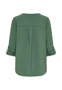 Blouse verte texturée avec des manches retroussées et un col arrondi. Le dos présente un détail de couture et un pli central pour un style supplémentaire.