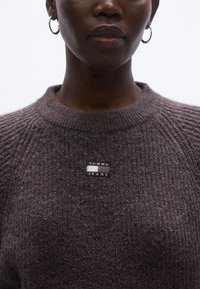 Pulover gri tricotat cu decolteu rotund și o etichetă cu logo-ul TOMMY JEANS pe față, având un material texturat și confortabil.