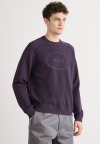Ljubičasti pleteni dukserica s okruglim izrezom, rebrastim rukavima i uvučenim "BOSS" logotipom u središtu. Upareno s sivim hlačama.