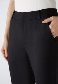 Pantalons noirs ajustés avec une texture lisse, dotés de poches latérales et d'une ceinture structurée. Vue rapprochée de la taille jusqu'au mi-thigh.