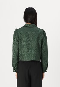 Grön kort jacka i texturerad tyg med utsmyckade mönster. Har en krage och knäppta manschetter, stylad med pösiga ärmar.