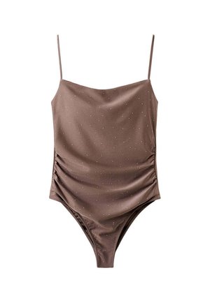 Donna con costume da bagno marrone e occhiali da sole in piedi scalza su una riva rocciosa con spiaggia sabbiosa e colline sullo sfondo.