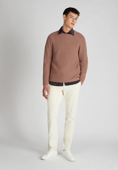 Brauner gerippter Pullover mit rundem Ausschnitt, kombiniert mit einem dunklen Hemd mit Kragen, hellen Cordhosen und weißen Sneakers.