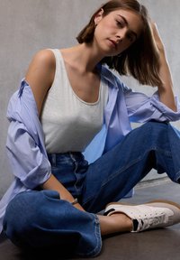 Top grigio sottostante una camicia a righe blu chiaro, abbinato a jeans in denim a vita alta e gamba larga, e sneakers bianche su uno sfondo di cemento.