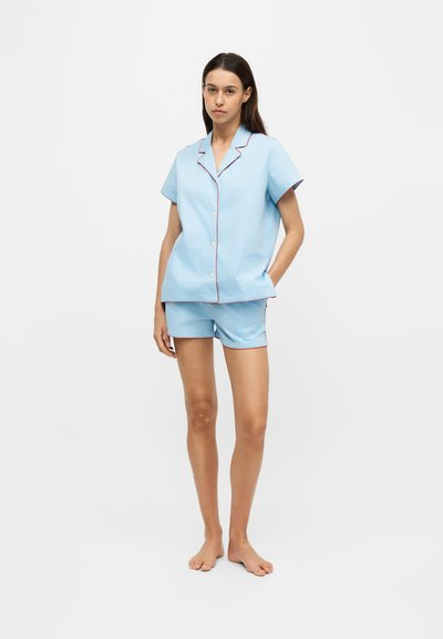 Femme debout pieds nus, portant un haut boutonné à manches courtes bleu clair et un short assorti avec bordure foncée sur un fond blanc uni.