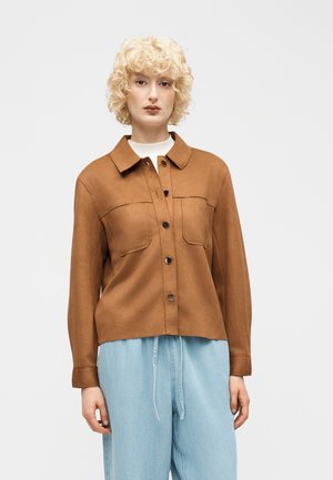 Femme blonde portant une veste marron à boutons avec deux poches poitrine et un jean bleu clair ample, debout devant un fond uni.