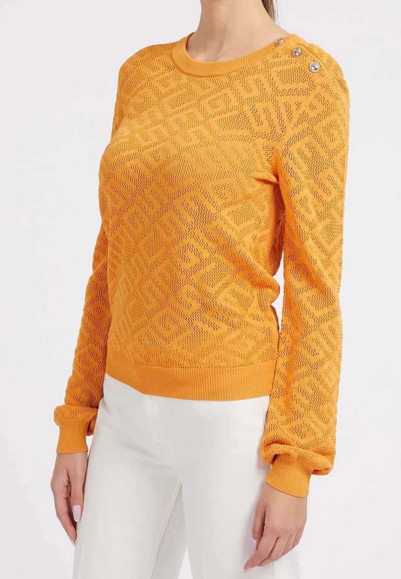 Femme portant un pull en maille orange ajusté et texturé avec des motifs géométriques et des détails de boutons sur l'épaule gauche, associé à un pantalon blanc.
