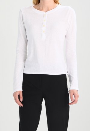 Mujer vestida con una camisa henley blanca de manga larga con botones y pantalones negros, de pie frente a un fondo liso.