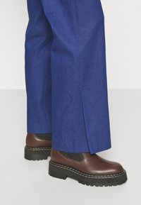 Bottes en cuir marron à chevilles avec des côtés élastiques noirs, dotées d'une épaisse semelle en crêpe noire et de coutures blanches, associées à un pantalon large bleu.