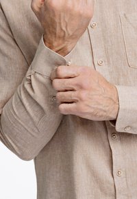 Camisa de lino beige con cuello abotonado. Presenta un bolsillo en el pecho y puños abotonados. Textura visible y un sutil tejido de tela.