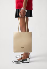 Beige lærveske med flate håndtak, glatt tekstur og preget logo. Modellen har på seg sølvfargede leiligheter og en rød genser.