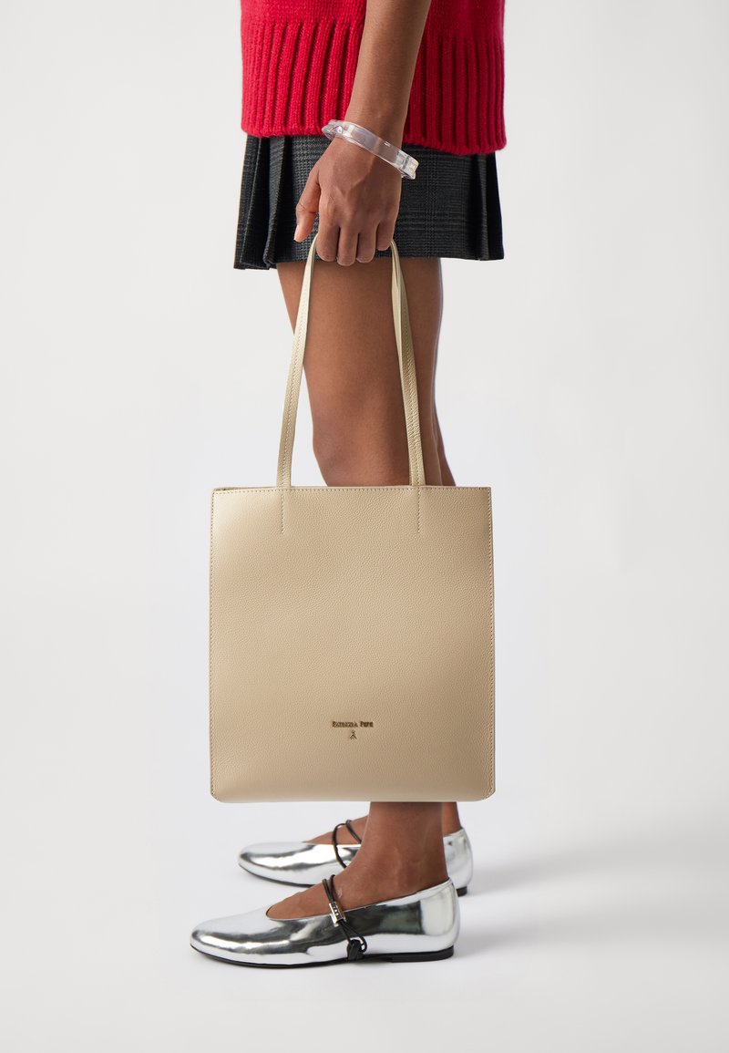 Beige lærveske med flate håndtak, glatt tekstur og preget logo. Modellen har på seg sølvfargede leiligheter og en rød genser.