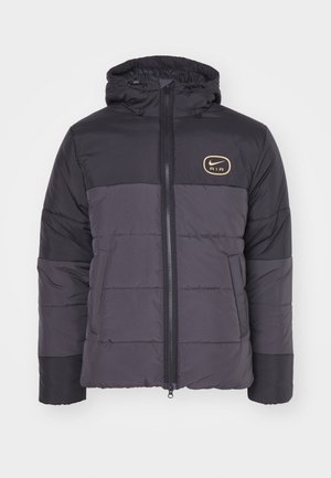 Giacca a vento nera e grigia con zip frontale, cappuccio regolabile, polsini a costine e logo dorato Nike Air sul petto. Dettaglio della cucitura orizzontale.