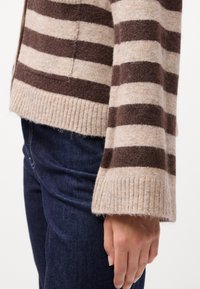 Ριγέ πλεκτό πουλόβερ σε μπεζ και καφέ αποχρώσεις, με ελαστικά μανίκια και μαλακή υφή, συνδυασμένο με σκούρα μπλε τζιν.