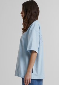 Camiseta de gran tamaño azul claro con mangas cortas, escote redondo y un pequeño detalle de logo en el lado. Tejido suave con una textura lisa.