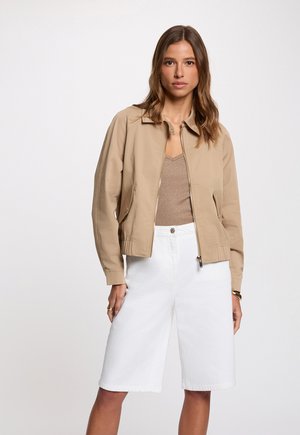 Femme portant une veste beige zippée par-dessus un top beige avec un short blanc ample au genou, debout devant un fond clair uni.