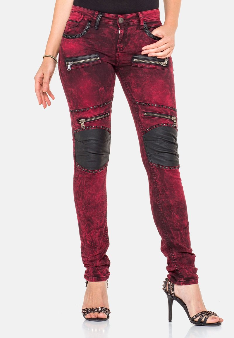 Femme portant un jean skinny rouge avec des empiècements noirs aux genoux, plusieurs zips, des détails à clous et des sandales noires à talons hauts, debout sur fond blanc.
