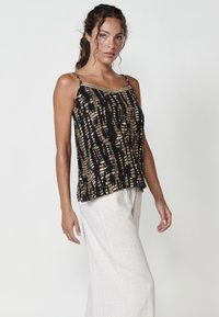 Zwarte tanktop met een gouden abstract patroon, dunne bandjes en een decoratieve rand langs de halslijn. Gecombineerd met lichtgekleurde broek.