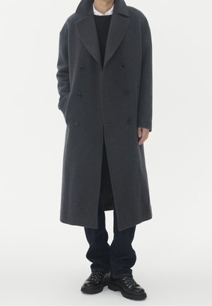 Person wearing a long dark gray double-breasted coat with wide lapels, dark pants, and black lace-up boots, standing with hands in pockets.