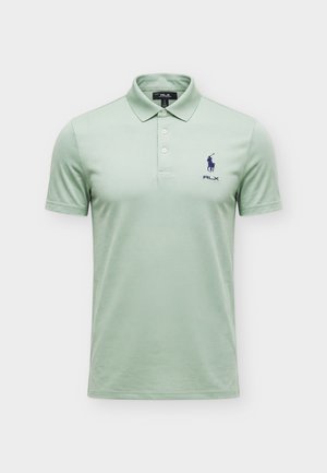 Hombre de pie mirando hacia adelante, con camiseta tipo polo verde claro, pantalones cortos negros con cinturón, calcetines blancos y zapatillas blancas sobre fondo liso.