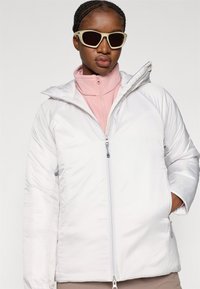 Veste blanche matelassée avec capuche, équipée d'une fermeture éclair complète et de poches latérales. Portée par-dessus un haut zippé rose clair. Intégrée dans un fond minimaliste.