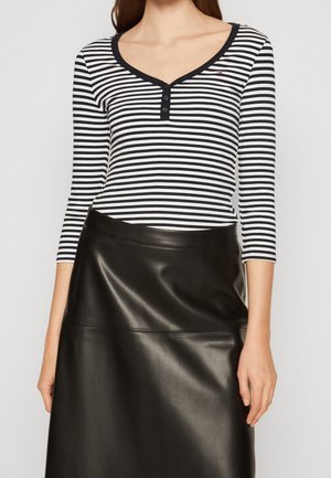 Femme portant un haut à manches longues rayé noir et blanc avec des boutons et une jupe en cuir noire, montrée du milieu de la poitrine jusqu'au milieu de la cuisse.