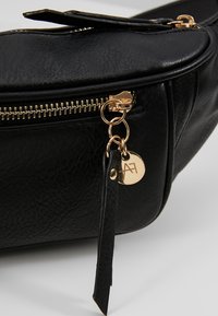 Zwarte faux leren tas met een gouden rits en hardware. Bevat een ronde metalen tag die is bevestigd met een leren riempje. Glad textuur.