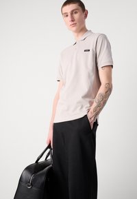Polo beige clair avec un logo noir, manches courtes et col, associé à un pantalon noir ample et un sac en cuir noir.