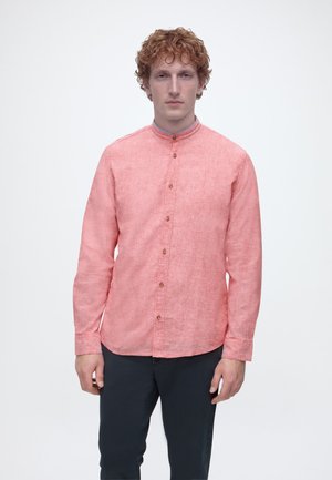 Muž s kučeravými červenými vlasmi, oblečený v koralovej košeli na gombíky s dlhými rukávmi a tmavých nohaviciach, stojaci na jednoduchom bielom pozadí.