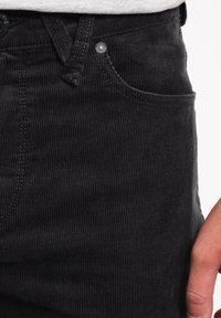 Pantalons en velours côtelé noir avec des côtes verticales, dotés d'une poche avant et d'une couture distinctive. Le tissu a une texture douce.