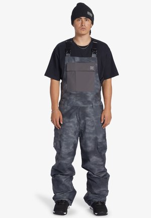 Grå snedriver med struktureret overflade, justerbare stropper, stor frontlomme og cargo-lommer, parret med en sort hue.
