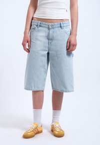 Shorts in denim azzurro chiaro con una vestibilità ampia, cinque tasche e orlo sfilacciato. Abbinati a sneakers gialle e calzini bianchi alla caviglia.