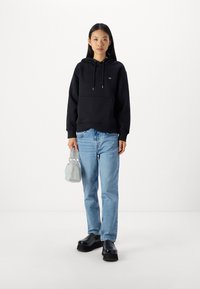 Felpa nera con un tascone frontale, dotata di cordini e un piccolo logo. Indossata con jeans in denim blu chiaro e scarpe nere chunky.