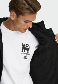 Zwarte ritssluitingjack over een wit T-shirt met een ontwerp van een zwarte kat en tekst. De stof lijkt glad met een moderne snit.