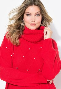 Roter gestrickter Pullover mit Rollkragen, der mit Rautenmustern und dekorativen weißen Perlen verziert ist. Gerippte Bündchen und Kragen sorgen für Textur.