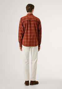 Camicia di flanella a quadri nei toni del rosso e dell'arancione, con colletto a camicia e maniche lunghe, abbinata a pantaloni bianchi e stivali marroni.